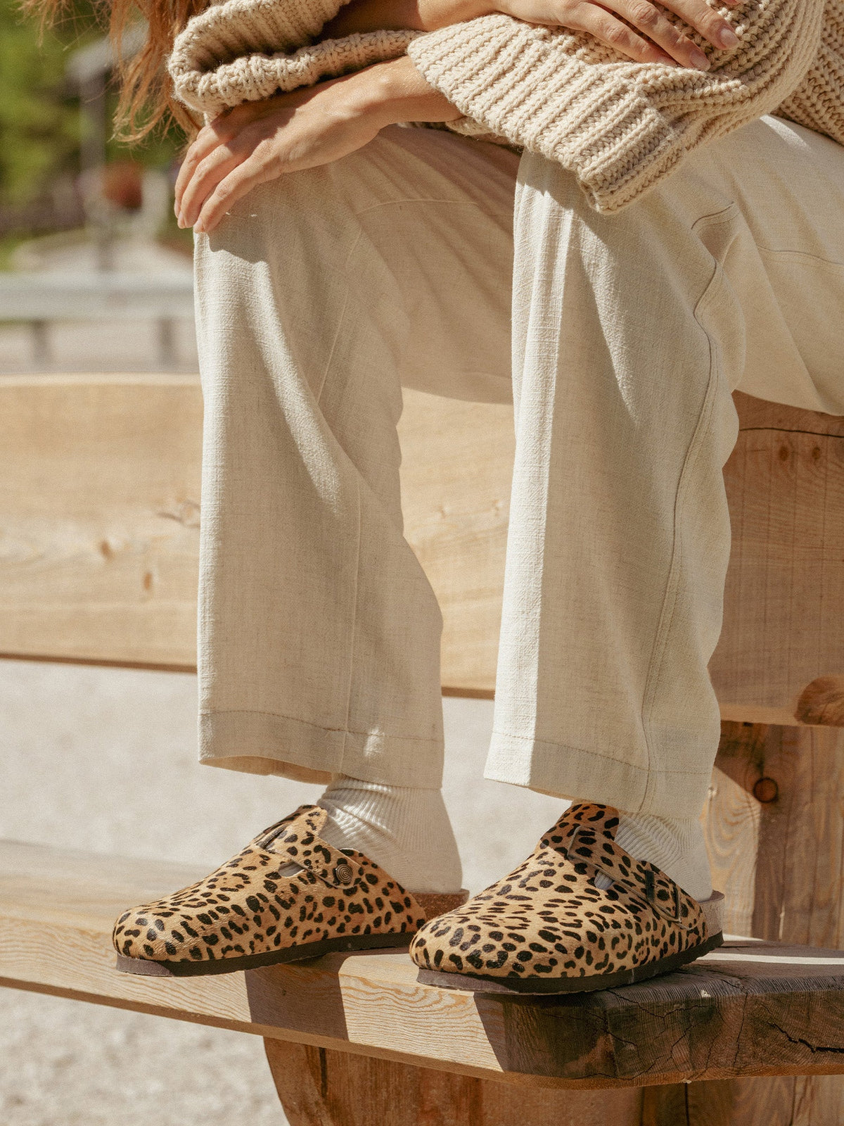 Close-up of Genuins Riva clogs in leopard print worn with cream trousers on a wooden bench